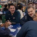 A group of military personnel in navy blue uniforms are gathered around a table in what appears to be a mess hall or dining facility aboard a naval vessel. Several sailors are engaged in conversation, smiling and interacting in a relaxed social setting. The background shows additional personnel seated at other tables in a busy dining environment.