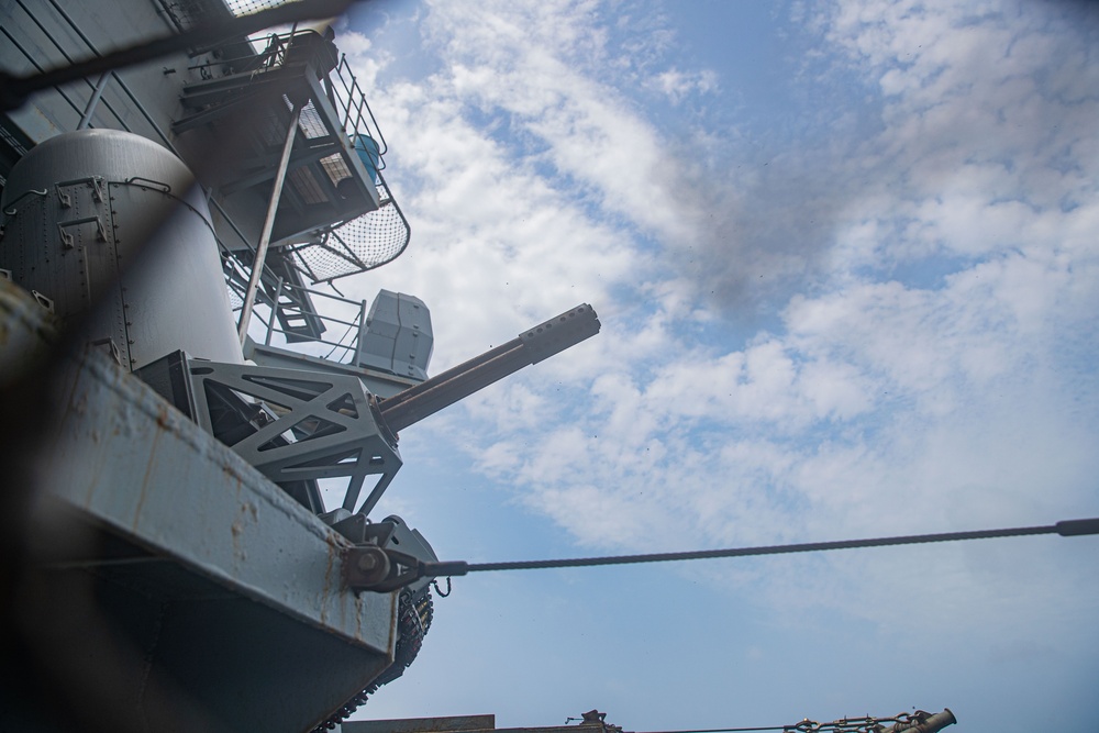 A low-angle upward shot taken aboard a naval vessel, showing a deck-mounted gun system elevated toward a partly cloudy sky. The image captures structural elements of the ship including superstructure, railings, ladders, and rigging. The perspective suggests the photo was taken from deck level looking upward.