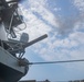 A low-angle upward shot taken aboard a naval vessel, showing a deck-mounted gun system elevated toward a partly cloudy sky. The image captures structural elements of the ship including superstructure, railings, ladders, and rigging. The perspective suggests the photo was taken from deck level looking upward.