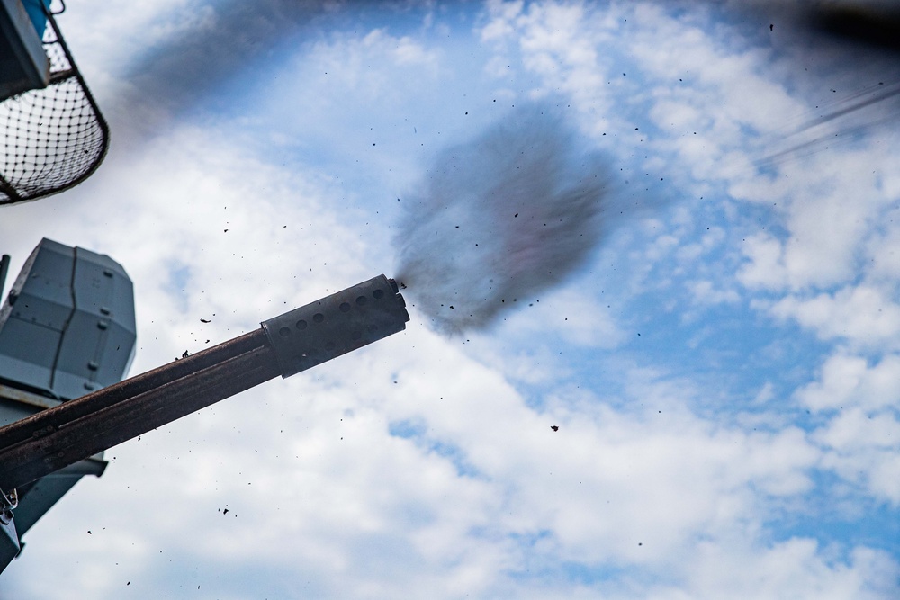The image captures a naval gun firing from a low angle perspective, with a visible muzzle blast and smoke cloud expanding into a partly cloudy sky. Debris and propellant particles are visibly scattered around the muzzle. The photograph appears to be taken from the deck of a warship, with gray naval superstructure visible on the left side.