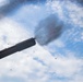 The image captures a naval gun firing from a low angle perspective, with a visible muzzle blast and smoke cloud expanding into a partly cloudy sky. Debris and propellant particles are visibly scattered around the muzzle. The photograph appears to be taken from the deck of a warship, with gray naval superstructure visible on the left side.