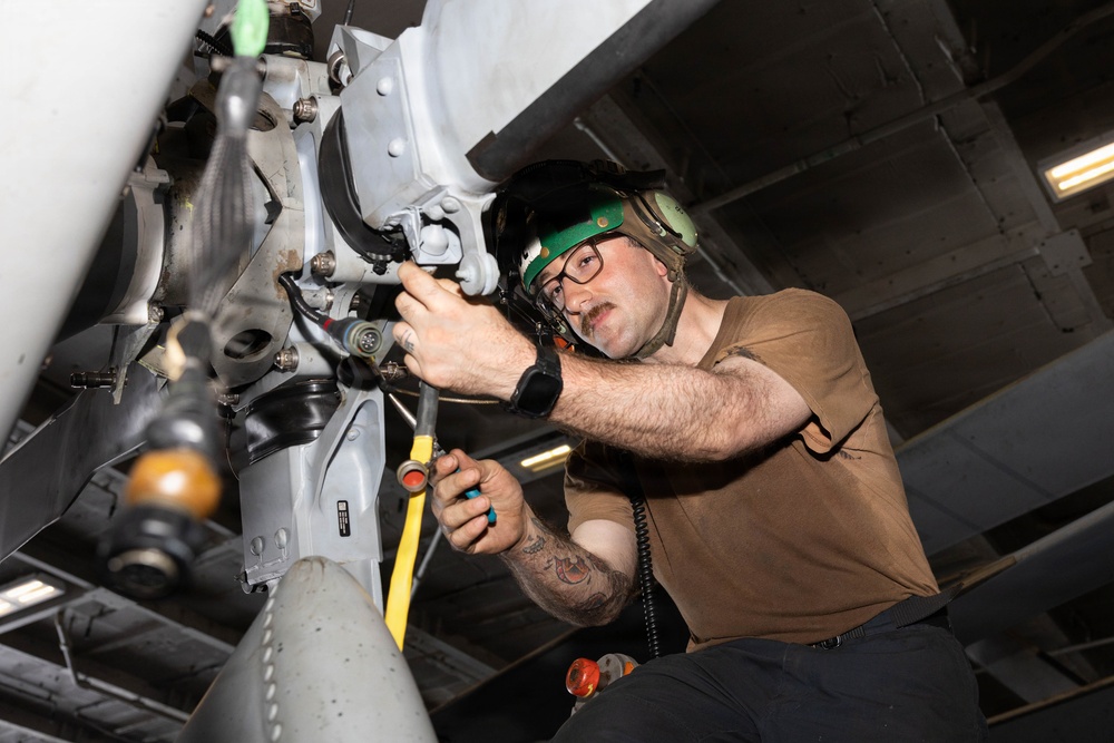 A military aviation technician is performing maintenance work on the underside of an aircraft, likely in a hangar bay or below-deck environment. The individual is using hand tools to work on what appears to be a weapons pylon or landing gear assembly. The worker is wearing standard military maintenance attire including hearing protection and safety glasses.