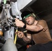 A military aviation technician is performing maintenance work on the underside of an aircraft, likely in a hangar bay or below-deck environment. The individual is using hand tools to work on what appears to be a weapons pylon or landing gear assembly. The worker is wearing standard military maintenance attire including hearing protection and safety glasses.