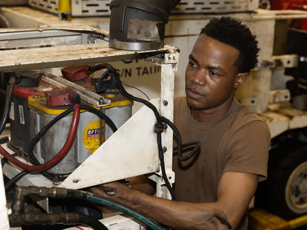 A young man in a brown t-shirt is working on mechanical or electrical equipment, closely inspecting a battery compartment area. The scene appears to be inside a maintenance bay or workshop environment. He is focused and engaged in what appears to be a repair or inspection task.