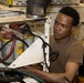 A young man in a brown t-shirt is working on mechanical or electrical equipment, closely inspecting a battery compartment area. The scene appears to be inside a maintenance bay or workshop environment. He is focused and engaged in what appears to be a repair or inspection task.