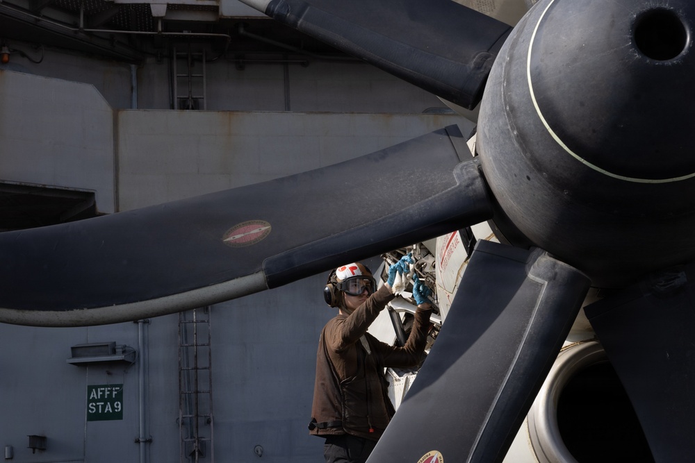 A naval aviation mechanic or deck crew member is performing maintenance on a large multi-blade propeller assembly, likely on an aircraft aboard a naval vessel. The worker is wearing standard flight deck safety gear including a helmet and protective goggles. A sign reading 'AFFF STA 9' is visible in the background, indicating an Aqueous Film Forming Foam fire suppression station aboard a ship.