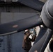 A naval aviation mechanic or deck crew member is performing maintenance on a large multi-blade propeller assembly, likely on an aircraft aboard a naval vessel. The worker is wearing standard flight deck safety gear including a helmet and protective goggles. A sign reading 'AFFF STA 9' is visible in the background, indicating an Aqueous Film Forming Foam fire suppression station aboard a ship.
