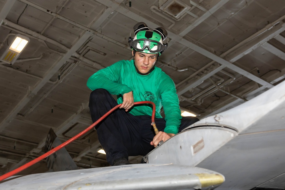 A military aviation technician wearing a green jersey and helmet with goggles is crouched on top of an aircraft, performing maintenance or fueling operations inside what appears to be a naval vessel's hangar bay. He is handling a red hose connected to a fitting on the aircraft's fuselage. The green jersey indicates his role as an aviation fuels or maintenance crew member aboard a US Navy aircraft carrier.