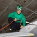 A military aviation technician wearing a green jersey and helmet with goggles is crouched on top of an aircraft, performing maintenance or fueling operations inside what appears to be a naval vessel's hangar bay. He is handling a red hose connected to a fitting on the aircraft's fuselage. The green jersey indicates his role as an aviation fuels or maintenance crew member aboard a US Navy aircraft carrier.