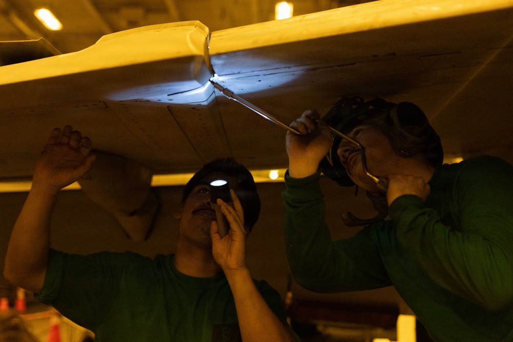 Two technicians in green shirts are working underneath what appears to be an aircraft wing or fuselage panel in a dimly lit hangar. One worker uses a headlamp or small flashlight to illuminate the work area while the other uses a tool to work on a joint or fastener point. The scene suggests maintenance, inspection, or repair work being performed on an aircraft structure.