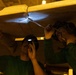Two technicians in green shirts are working underneath what appears to be an aircraft wing or fuselage panel in a dimly lit hangar. One worker uses a headlamp or small flashlight to illuminate the work area while the other uses a tool to work on a joint or fastener point. The scene suggests maintenance, inspection, or repair work being performed on an aircraft structure.