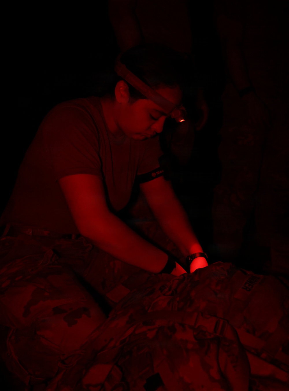 A person wearing a headlamp and a wristband is working in near-total darkness, illuminated entirely by red light. They appear to be performing a hands-on task over what looks like camouflage-patterned material, possibly conducting a medical or tactical procedure. The red lighting suggests a low-visibility operational or training environment.