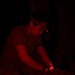 A person wearing a headlamp and a wristband is working in near-total darkness, illuminated entirely by red light. They appear to be performing a hands-on task over what looks like camouflage-patterned material, possibly conducting a medical or tactical procedure. The red lighting suggests a low-visibility operational or training environment.