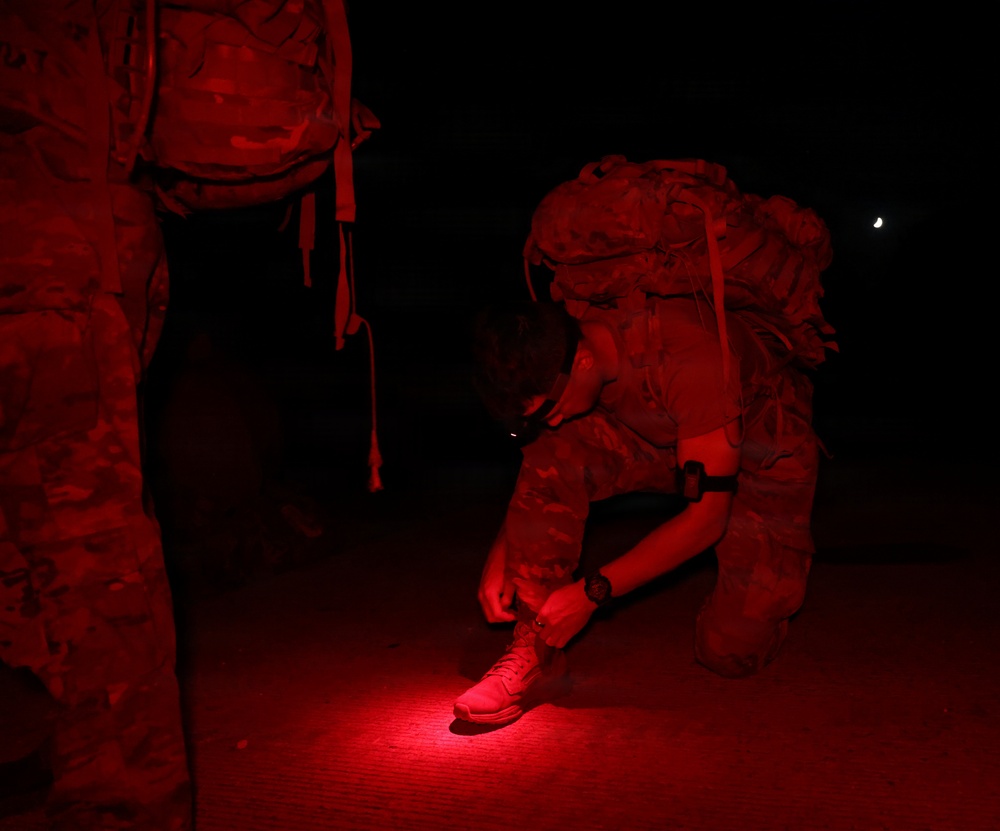 A military personnel in camouflage uniform and full pack is crouched down at night, using a red-light flashlight to adjust or tie their boot. A second uniformed individual stands nearby on the left. The scene is illuminated almost entirely by red light, consistent with tactical night operations where red light preserves night vision.