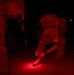 A military personnel in camouflage uniform and full pack is crouched down at night, using a red-light flashlight to adjust or tie their boot. A second uniformed individual stands nearby on the left. The scene is illuminated almost entirely by red light, consistent with tactical night operations where red light preserves night vision.