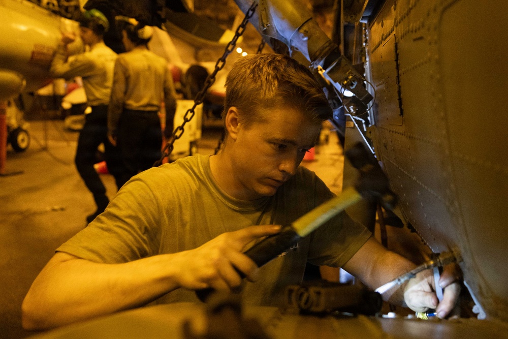 A young male technician or military maintainer is performing hands-on maintenance work on the fuselage of a military aircraft, likely in a carrier hangar bay given the low amber lighting and chain tie-downs visible in the background. He is using tools and a flashlight to work on a panel or component on the aircraft's skin. Additional personnel and aircraft are visible in the dimly lit background.
