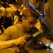 A young male technician or military maintainer is performing hands-on maintenance work on the fuselage of a military aircraft, likely in a carrier hangar bay given the low amber lighting and chain tie-downs visible in the background. He is using tools and a flashlight to work on a panel or component on the aircraft's skin. Additional personnel and aircraft are visible in the dimly lit background.