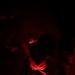 A person in military/tactical camouflage gear is illuminated by red light in an otherwise dark environment. They appear to be handling straps or equipment, possibly adjusting or packing tactical gear. The red lighting is consistent with night operations protocols, as red light preserves night vision.