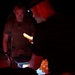 A nighttime scene showing military personnel gathered around a table or supply station illuminated by red light and a handheld light source. One individual in military tactical gear with a backpack and headlamp is examining items on the surface. The low-light conditions suggest a field operation or nighttime logistics activity.