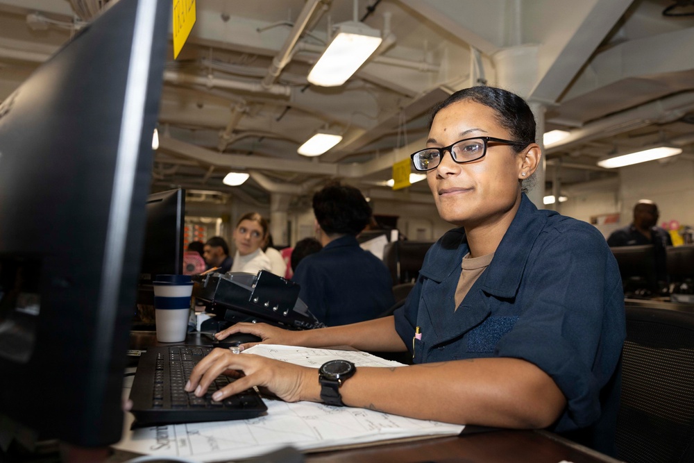 A woman wearing glasses and a dark navy blue uniform is seated at a workstation, typing on a keyboard while looking toward the camera. The setting appears to be a military or government office environment, likely aboard a naval vessel based on the industrial ceiling infrastructure and workspace layout. Several other personnel are visible working in the background.