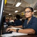 A woman wearing glasses and a dark navy blue uniform is seated at a workstation, typing on a keyboard while looking toward the camera. The setting appears to be a military or government office environment, likely aboard a naval vessel based on the industrial ceiling infrastructure and workspace layout. Several other personnel are visible working in the background.