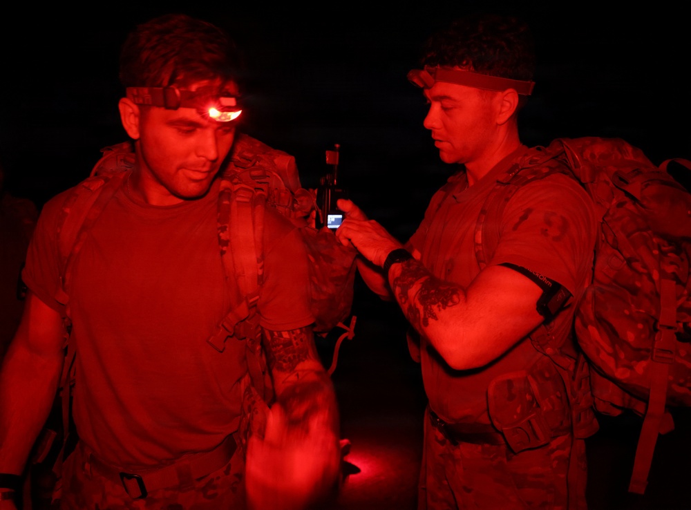 Two military personnel are photographed at night under red light illumination, both wearing tactical gear including backpacks and headlamps. One individual has an active red headlamp and the other appears to be checking a small device, possibly a phone or GPS unit. The scene suggests a nighttime military training exercise or operational activity.