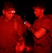 Two military personnel are photographed at night under red light illumination, both wearing tactical gear including backpacks and headlamps. One individual has an active red headlamp and the other appears to be checking a small device, possibly a phone or GPS unit. The scene suggests a nighttime military training exercise or operational activity.