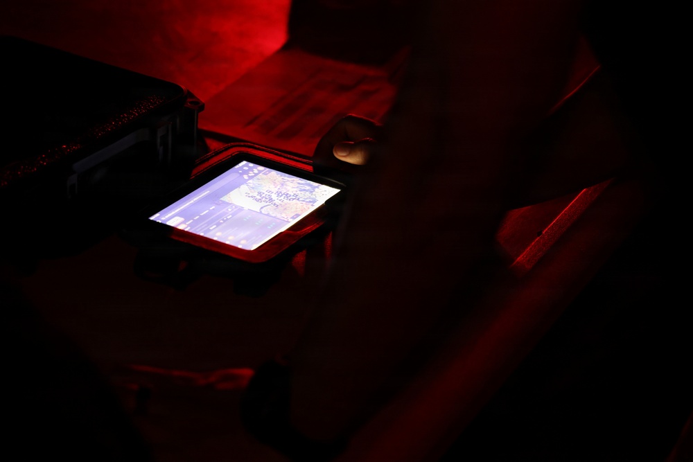 A person is viewing a small illuminated screen displaying what appears to be a digital map or navigation interface in a very dark, red-lit environment. The device appears to be housed in or placed on a rugged protective case. The scene suggests a tactical, operational, or field setting based on the lighting conditions and equipment visible.