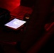 A person is viewing a small illuminated screen displaying what appears to be a digital map or navigation interface in a very dark, red-lit environment. The device appears to be housed in or placed on a rugged protective case. The scene suggests a tactical, operational, or field setting based on the lighting conditions and equipment visible.