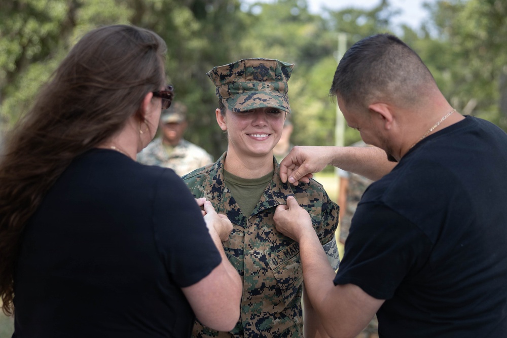 A U.S. Marine in woodland MARPAT camouflage uniform is smiling while two civilians in black shirts pin insignia or rank devices onto her collar. This appears to be a military promotion or pinning ceremony conducted outdoors. The Marine looks proud and happy during what is likely a significant career milestone.