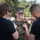 A U.S. Marine in woodland MARPAT camouflage uniform is smiling while two civilians in black shirts pin insignia or rank devices onto her collar. This appears to be a military promotion or pinning ceremony conducted outdoors. The Marine looks proud and happy during what is likely a significant career milestone.