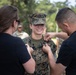 A U.S. Marine in woodland MARPAT camouflage uniform is smiling while two civilians in black shirts pin insignia or rank devices onto her collar. This appears to be a military promotion or pinning ceremony conducted outdoors. The Marine looks proud and happy during what is likely a significant career milestone.