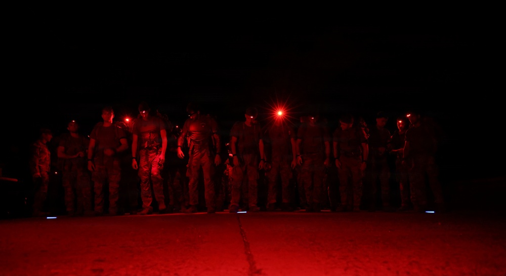 A group of military or tactical personnel are standing in a line formation at night, illuminated by red light sources. Several individuals are wearing red headlamps or helmet-mounted lights, casting a deep crimson glow across the scene. The setting appears to be an outdoor operational area, possibly a tarmac or staging ground.