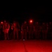 A group of military or tactical personnel are standing in a line formation at night, illuminated by red light sources. Several individuals are wearing red headlamps or helmet-mounted lights, casting a deep crimson glow across the scene. The setting appears to be an outdoor operational area, possibly a tarmac or staging ground.