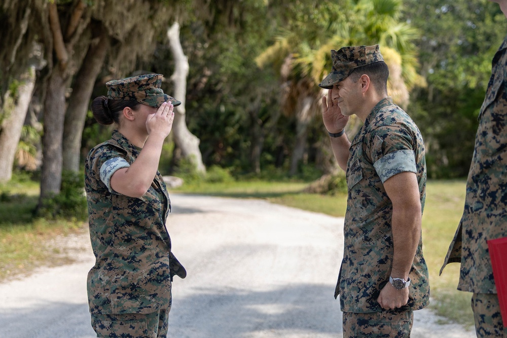 Two U.S. Marines in MARPAT woodland camouflage uniforms are exchanging salutes on a paved path in an outdoor setting with trees and vegetation in the background. A female Marine on the left is saluting a male Marine on the right, suggesting a formal military greeting or acknowledgment of rank. A third uniformed individual is partially visible on the far right.