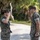 Two U.S. Marines in MARPAT woodland camouflage uniforms are exchanging salutes on a paved path in an outdoor setting with trees and vegetation in the background. A female Marine on the left is saluting a male Marine on the right, suggesting a formal military greeting or acknowledgment of rank. A third uniformed individual is partially visible on the far right.