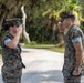 Two U.S. Marines in MARPAT woodland camouflage uniforms are exchanging salutes on a paved path in an outdoor setting with trees and vegetation in the background. A female Marine on the left is saluting a male Marine on the right, suggesting a formal military greeting or acknowledgment of rank. A third uniformed individual is partially visible on the far right.