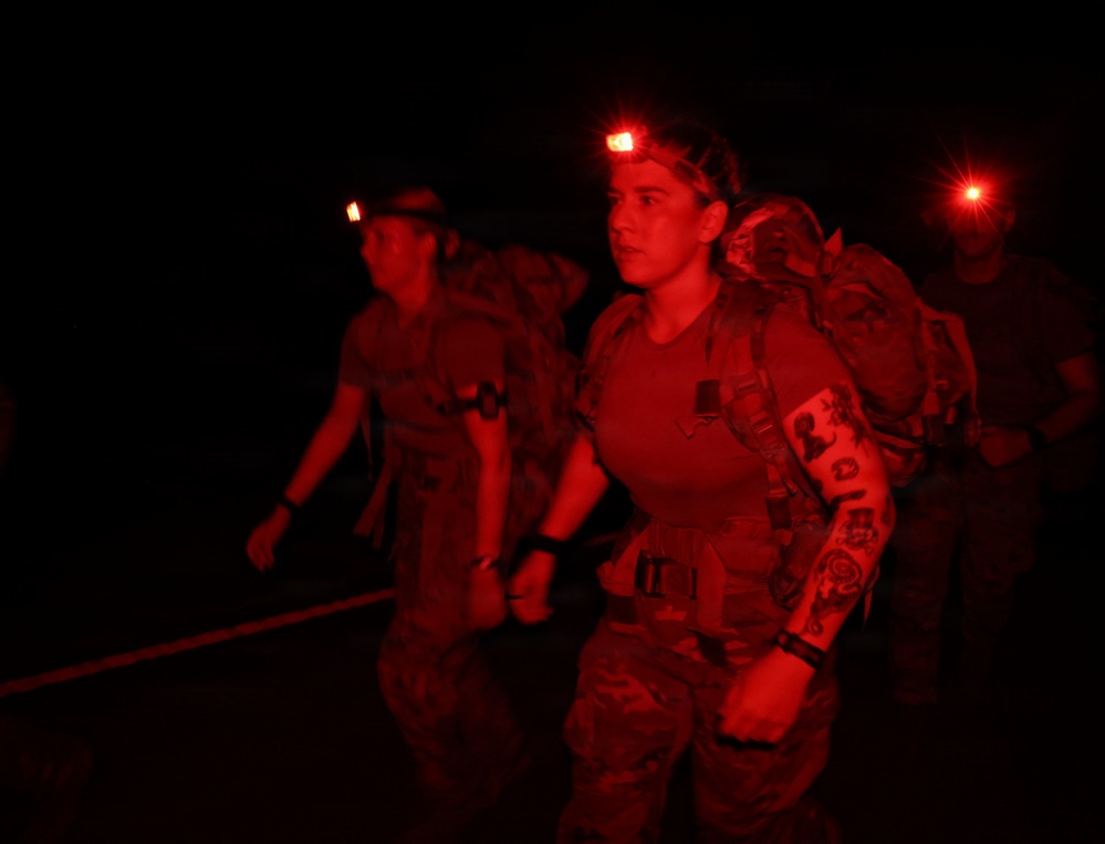 A group of military personnel conducting a nighttime ruck march or training exercise, illuminated by red-light headlamps. The foreground figure is a female service member wearing camouflage uniform, carrying a loaded rucksack, and displaying visible tattoos on her right arm. The red lighting suggests use of tactical night-vision-compatible illumination to preserve low visibility.