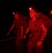 A group of military personnel conducting a nighttime ruck march or training exercise, illuminated by red-light headlamps. The foreground figure is a female service member wearing camouflage uniform, carrying a loaded rucksack, and displaying visible tattoos on her right arm. The red lighting suggests use of tactical night-vision-compatible illumination to preserve low visibility.