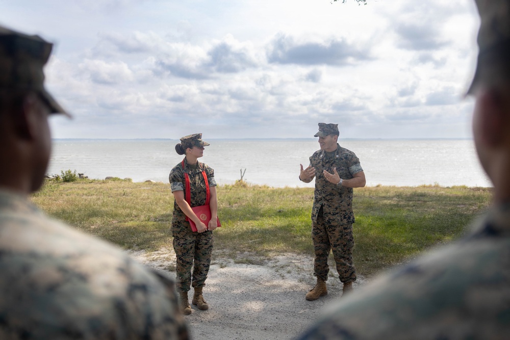 Two U.S. Marines in woodland MARPAT camouflage uniforms are engaged in conversation near a waterfront location, with additional Marines visible in the foreground as blurred figures. The female Marine is holding a red folder/binder while the male Marine appears to be briefing or instructing. The setting appears to be an outdoor coastal or lakeside area during daytime.