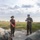 Two U.S. Marines in woodland MARPAT camouflage uniforms are engaged in conversation near a waterfront location, with additional Marines visible in the foreground as blurred figures. The female Marine is holding a red folder/binder while the male Marine appears to be briefing or instructing. The setting appears to be an outdoor coastal or lakeside area during daytime.