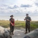 Two U.S. Marines in woodland MARPAT camouflage uniforms are engaged in conversation near a waterfront location, with additional Marines visible in the foreground as blurred figures. The female Marine is holding a red folder/binder while the male Marine appears to be briefing or instructing. The setting appears to be an outdoor coastal or lakeside area during daytime.