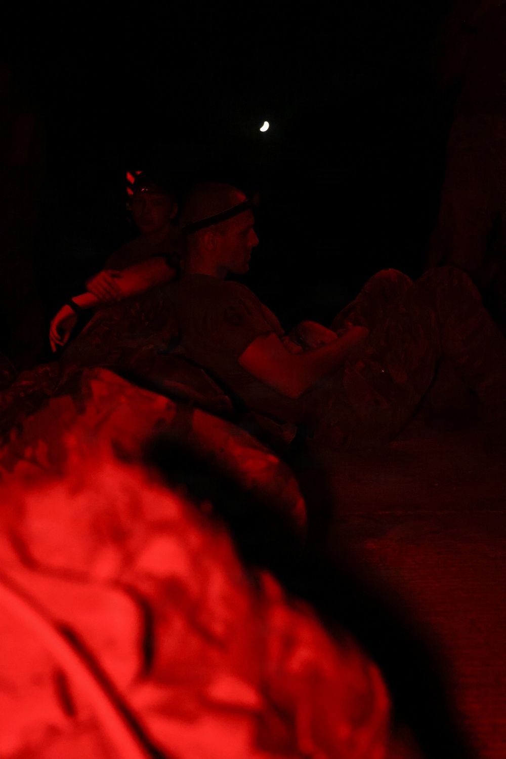 A nighttime scene illuminated by red light showing what appears to be military personnel wearing headlamps and camouflage gear. A crescent moon is visible in the dark sky above. The foreground contains a blurred, brightly red-lit object, likely military equipment or gear.