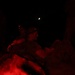 A nighttime scene illuminated by red light showing what appears to be military personnel wearing headlamps and camouflage gear. A crescent moon is visible in the dark sky above. The foreground contains a blurred, brightly red-lit object, likely military equipment or gear.