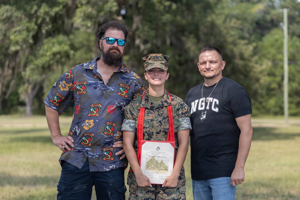 Three individuals pose together outdoors, with the person in the center wearing United States Marine Corps camouflage uniform and cover (cap), holding a framed United States Marine Corps certificate with a red ribbon. The two individuals flanking the Marine appear to be family members or supporters, dressed in civilian clothing. The setting appears to be a grassy outdoor area, likely during a military ceremony or recognition event.