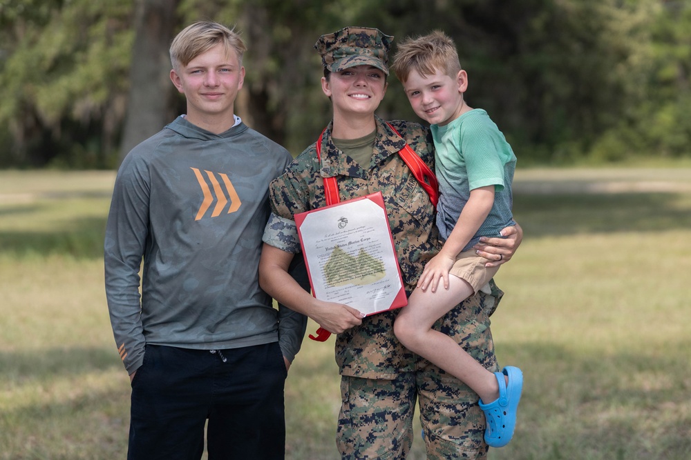 A U.S. Marine in digital camouflage uniform holds a United States Marine Corps certificate while posing with two children in an outdoor grassy setting. She is holding a young boy on her hip while an older teenage boy stands to her left. The image appears to capture a military achievement or promotion ceremony moment with family present.