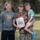 A U.S. Marine in digital camouflage uniform holds a United States Marine Corps certificate while posing with two children in an outdoor grassy setting. She is holding a young boy on her hip while an older teenage boy stands to her left. The image appears to capture a military achievement or promotion ceremony moment with family present.