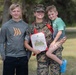 A U.S. Marine in digital camouflage uniform holds a United States Marine Corps certificate while posing with two children in an outdoor grassy setting. She is holding a young boy on her hip while an older teenage boy stands to her left. The image appears to capture a military achievement or promotion ceremony moment with family present.