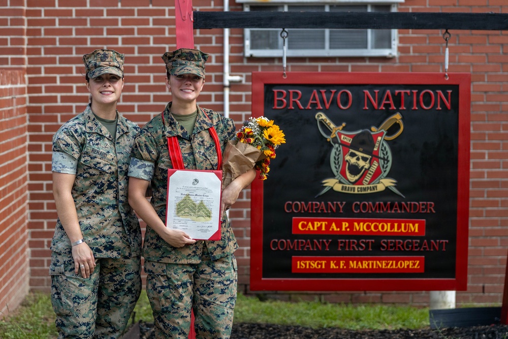 Two female U.S. Marines in MARPAT camouflage uniforms pose together in front of a 'Bravo Nation' company sign. One Marine is holding a bouquet of flowers and a framed certificate with a red ribbon, suggesting a recognition or promotion ceremony. The sign identifies the unit's Company Commander as Capt A.P. McCollum and Company First Sergeant as 1stSgt K.F. Martinezlopez.