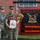 Two female U.S. Marines in MARPAT camouflage uniforms pose together in front of a 'Bravo Nation' company sign. One Marine is holding a bouquet of flowers and a framed certificate with a red ribbon, suggesting a recognition or promotion ceremony. The sign identifies the unit's Company Commander as Capt A.P. McCollum and Company First Sergeant as 1stSgt K.F. Martinezlopez.